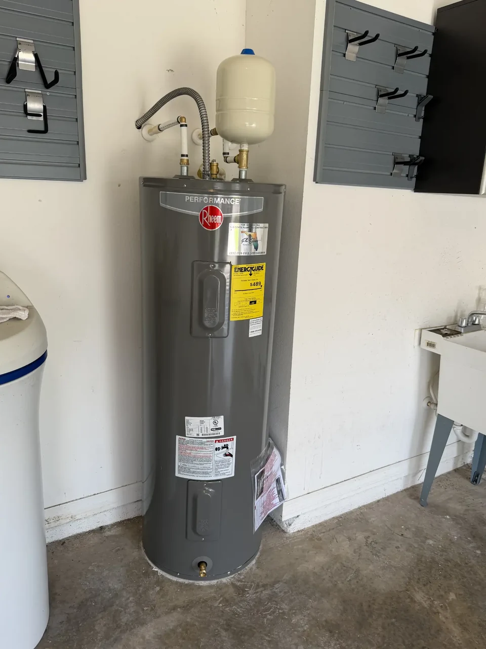 An electric tank water heater with an expansion tank and connected supply lines in a garage.