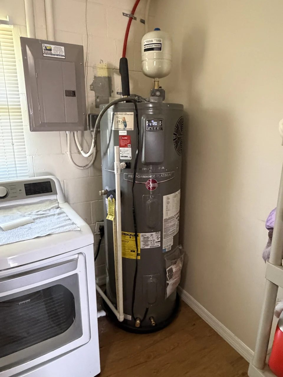 A Rheem hybrid heat pump water heater installed in a laundry room with an expansion tank above and connected pipework.